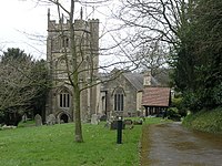 Square grey tower of stone church building, partially obscured by trees. Red roofed lych gate to right. Grass and gravestones in the foreground