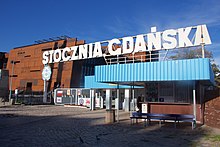 Biały szyld „Stocznia Gdańska” nad bramą nr 2 przed Europejskim Centrum Solidarności. Pod szyldem znajduje się ozdobna niebieska przesłona na styku dachu i ściany. Na bramie powieszony jest plakat z czerwonym napisem „Solidarność”, zdjęcie papieża Jana Pawła 2, obraz Matki Boskiej Częstochowskiej i flaga Polski. Za bramą budynek Europejskiego Centrum Solidarności w kolorze rdzy. W tle błękitne niebo.