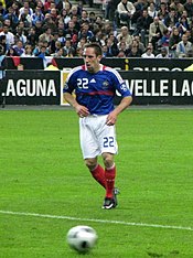 A young man with brown hair in red football kit jogging alongside the touch-line of a football pitch