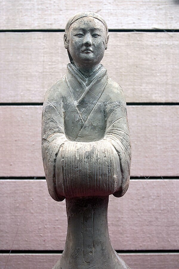 A Han Dynasty (202 BC – 220 AD) Chinese ceramic figurine of a lady's maid in a standard formal pose with hands covered by long sleeve cuffs in the tra