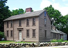 The main part of the home is a wooden, two-and-a-half story rectangular building with large windows, one central door, and a central chimney. A smaller wing extends back from the right side. There are large trees in the background and a low rock wall in the foreground.