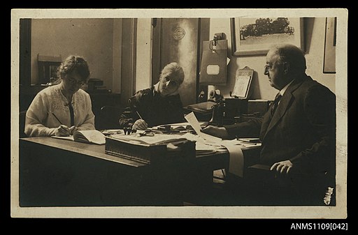 Photographic postcard of a man and two women seated at partners desk (10413330674)