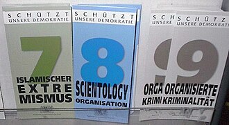 Display with three types of information leaflets, with the numbers and titles "7 Islamischer Extremismus" (coloured in shades of green), "8 Scientology Organisation" (coloured in shades of blue) and "9 Organisiserte Kriminalität" (shades of grey). At the top of each leaflet is the inscription "Schützt unsere Demokratie".