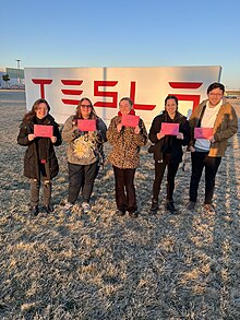 Five smiling workers are standing together. They are holding signs that say "union" wrapped in hearts. Behind them, is a concrete stand emblazoned with the Tesla logo.