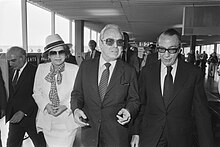 Photograph of a crowd walking through an airport terminal. In the front is a woman in a hat and two men in business suits.