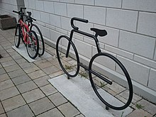 Dois bicicletários em forma de bicicletas estilizadas, em metal com acabamento em preto brilhante, cada um encaixado em um retângulo de concreto dentro de um piso de pequenos ladrilhos de cerâmica, em frente a uma parede de blocos de concreto pintada de branco.  O que está ao fundo tem uma mountain bike acorrentada.