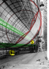 Looking down the interior of a partially completed airship frame. Two passageways are highlighted; one along the bottom and one right through the middle. The closest two structural polygons are also highlighted; the cantilevers on the nearest one are visible. The two people in the foreground are barely visible.