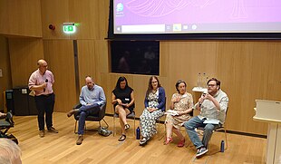 Five people seated, one speaking into a microphone. A sixth person is stood to the side holding another microphone.