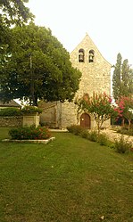 Vue de l'église à une seule nef, en pierre calcaire blanche et toit en tuiles plates, pas de clocher individualisé, place et calvaire devant