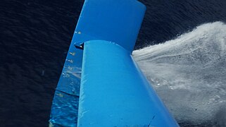 Segelboot Eraclide: Blick rechts des Rumpfs von oben auf die teilweise aus dem Wasser ragende V-Fläche des (nach links) reisenden Boots.