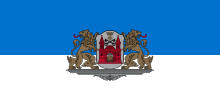 Eine blau-weiße Flagge mit einem Wappen in der Mitte. Das Wappen hat eine rote Burg mit einem Löwenkopf in dessen Zentrum sowie eine Krone, ein Kreuz und zwei überkreuzte Schlüssel. Das Wappenschild wird von zwei braunen Löwen gehalten, die ihre Köpfe nach außen gedreht haben und ihre Zunge ausstrecken. Wappen und Löwen stehen auf einem grauen Podest.