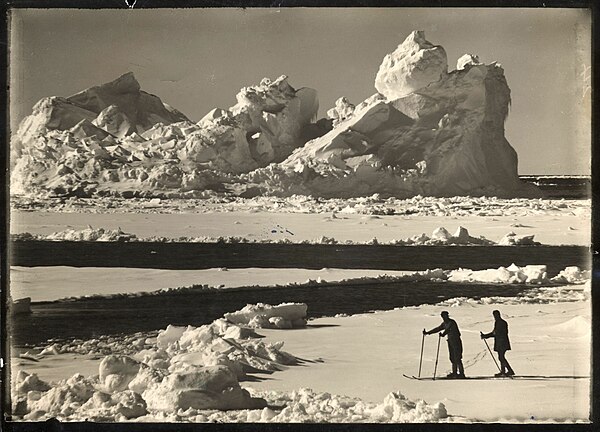 Photo of an eroded iceberg with two men skiing on an ice floe in the foreground