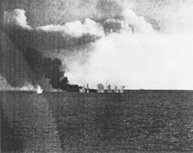USS Gambier Bay, burning from earlier gunfire damage, is bracketed by a salvo from a Japanese heavy cruiser (faintly visible in the background, center