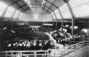 Darkened interior of a large hall with two rows of high windows along each side. It is possible to discern a seated orchestra in the foreground, with mass choirs in the background.