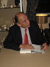 Een man met een rode stropdas, een geruit overhemd en een donkere jas die een boek signeert