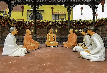 Sculptures of five people clad in traditional Indian clothing sitting under an ornate black canopy in a semicircle on the floor around another bespectacled person in saffron clothes.