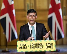 Sunak speaking, pen in hand, at a lectern with "Stay Alert > Control the Virus > Save Lives" on it, in a wood-panelled room with two Union flags in the background