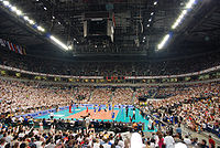 Mecz siatkówki w całkowicie wypełnionej kibicami Belgradzkiej Arenie. Kibice w większości ubrani są na biało. W lewym górnym rogu częściowo widoczne flagi pańśtw uczestniczących w turnieju. Zdjęcie wykonane z perspektywy trybun.