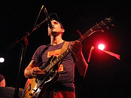 A worm's eye view shot of Sufjan Stevens playing an orange and black electric guitar while singing into a microphone. He is wearing a blue T-shirt with an orange letter "I" on it and orange pants.