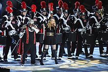 A groupe of performers onstage, with Madonna and Cee Lo Green at the front. They are all wearing black costumes with red and white stripes.