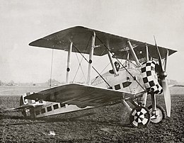 Three quarters view of military biplane on the ground with pilot in cockpit and check paint pattern on engine cowling and wheels