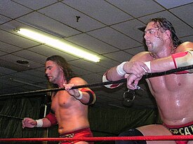 Dos hombres blancos vestidos de rojo equipo de lucha de pie en un ring de lucha con cuerdas rojas y negras.