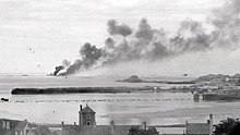 Photographie en noir et blanc montrant en premier plan le toit d'habitations (les bas Sablons), puis la mer, un môle, à nouveau la mer et en fond, une île d'où s'échappe une épaisse fumée. À droite, deux presqu'îles sont visibles (le petit Bé et le Grand Bé).