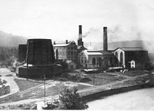 Fotografía en blanco y negro que muestra altos edificios industriales con dos torres de refrigeración de metal y dos chimeneas de fábrica.