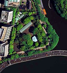 Kleurenfoto: luchtfoto van een bosrijke omgeving, waaruit enkele gebouwen verrijzen (een daarvan heeft een zeshoekig dak), aan de rand van een beek.
