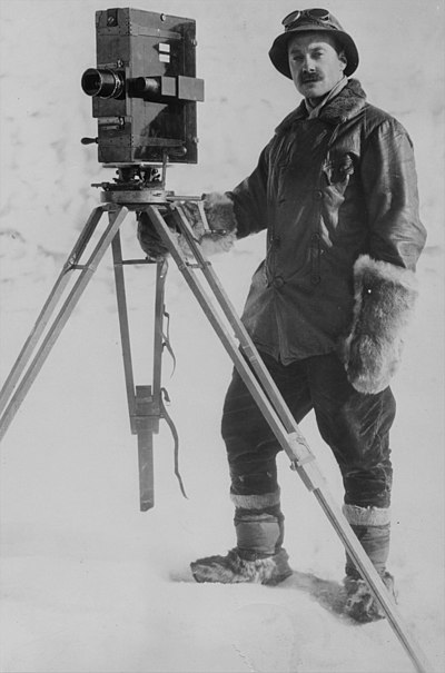 Photo of a man with thick mittens standing by a boxy camera on a tripod