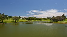 Photographie d'une partie d'un terrain de golf qui présente une grande étendue d'eau et une dense végétation tropicale.