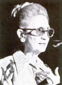 Seorang wanita tua dalam cahaya keras, abu-abu rambut updo bob, mengenakan kacamata berwarna dan berbicara pada mikrofon