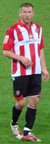 A man with short brown hair is wearing a red and white striped top, black shorts and black socks. He is standing on a grass field.