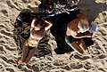 Topless couple on an Ajaccio beach, from above. The woman is lying down, but has her head and legs raised, so her face is not clearly visible. Her chest and thighs, including her nipples, are clearly visible. The man is sitting and reading a book; his face is not visible at all.