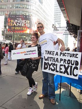 A girl holding a sign that says "LD = equally intelligent / Cross out stigma" poses for a photo in Times Square with a man holding a sign that says "Take a picture with a proud Dyslexic".