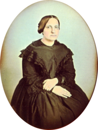 Photographic portrait of a woman seated and wearing a dark dress trimmed in dark lace, with her hair pulled back into a bun and no jewelry except for a simple ring on her left hand
