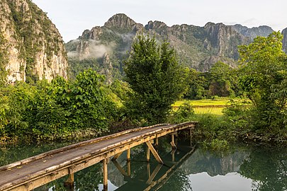 Three-quarter view of a wooden footbridge over a lagoon, trees and mountains in Vang Vieng, Laos