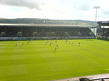 Een gras voetbalveld met markeringen erop geschilderd.  Achter het veld staat een overdekte tribune met houten stoelen en in de rechterbovenhoek staat een schijnwerpermast.