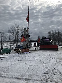 Mohawk-Demonstranten stehen neben einem mit Irokesen- und Mohawk-Krieger-Flaggen bedeckten Schneepflug in der Nähe eines Bahnübergangs in der Wyman Rd mit Brennholz und einem Holzschild mit der Aufschrift "#RCMP raus".