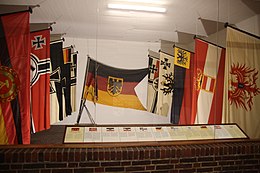Links und rechts je 5 Flaggen an Stöcken, hinten an der Wand die aktuelle Dienstflagge der Deutschen Marine, vorne Tafeln mit Erläuterungen zu jeder Flagge