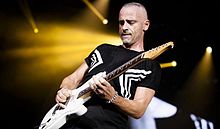 A man with short gray hair is wearing a black shirt and is playing on a white guitar