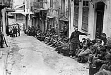 photographie noir et blanc : des hommes assis le long de maisons dans une rue en pente