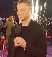 Grzegorz Damięcki mengenakan jas, memegang mikrofon, di premiere film Podatek od miłości di 2018