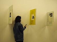 Una mujer con un teléfono inteligente frente a 3 paneles de vidrio perpendiculares a una pared.