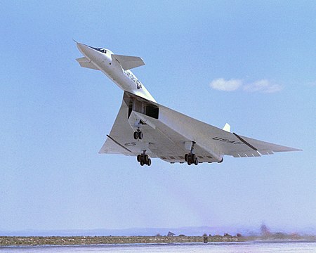 White delta-wing aircraft taking off with landing gears retracting. At the front of aircraft are canards.