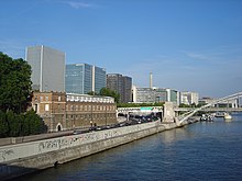 Seitliche Farbfotografie eines Ufers mit dem Fluss und einer Bogenbrücke aus Eisen auf der rechten Seite. Am Ufer ist eine Betonmauer mit einer befahrenen Straße darauf und moderne Bürogebäude.