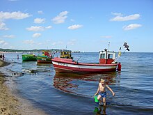 Zdjęcie trzech kutrów rybackich, cumujących na morzu przy plaży, oznaczonych literami „SOP” (czyli Sopot). Większą część zdjecia zajmuje morze, w tle widać molo w Sopocie oraz klif w Orłowie. Na pierwszym planie bawiący się w morzu chłopczyk z wiaderkiem.