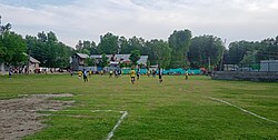 "Sports enthusiasts playing football on the lush green playground of Paribal Shallabugh, located on the bank of the Sind River"