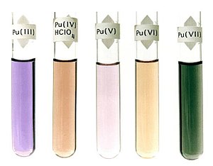 Five fluids in glass test tubes: violet, Pu(III); dark brown, Pu(IV)HClO4; light purple, Pu(V); light brown, Pu(VI); dark green, Pu(VII)