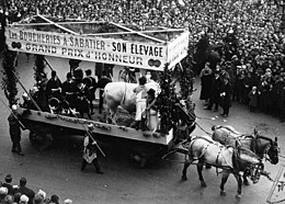 Photo en noir et blanc représentant un bœuf sur un char tiré par des chevaux et passant au milieu de la foule